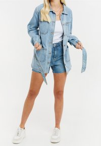 Chaqueta de mezclilla azul claro con botón frontal, dos bolsillos en el pecho y mangas largas. Combinada con pantalones cortos de mezclilla azul claro y zapatillas blancas.