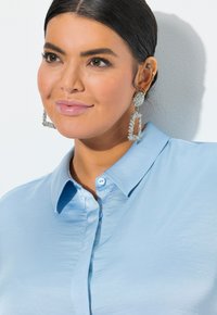 Camicia azzurro chiaro con texture liscia e colletto. Accompagnata da grandi orecchini statement in argento con design geometrico.