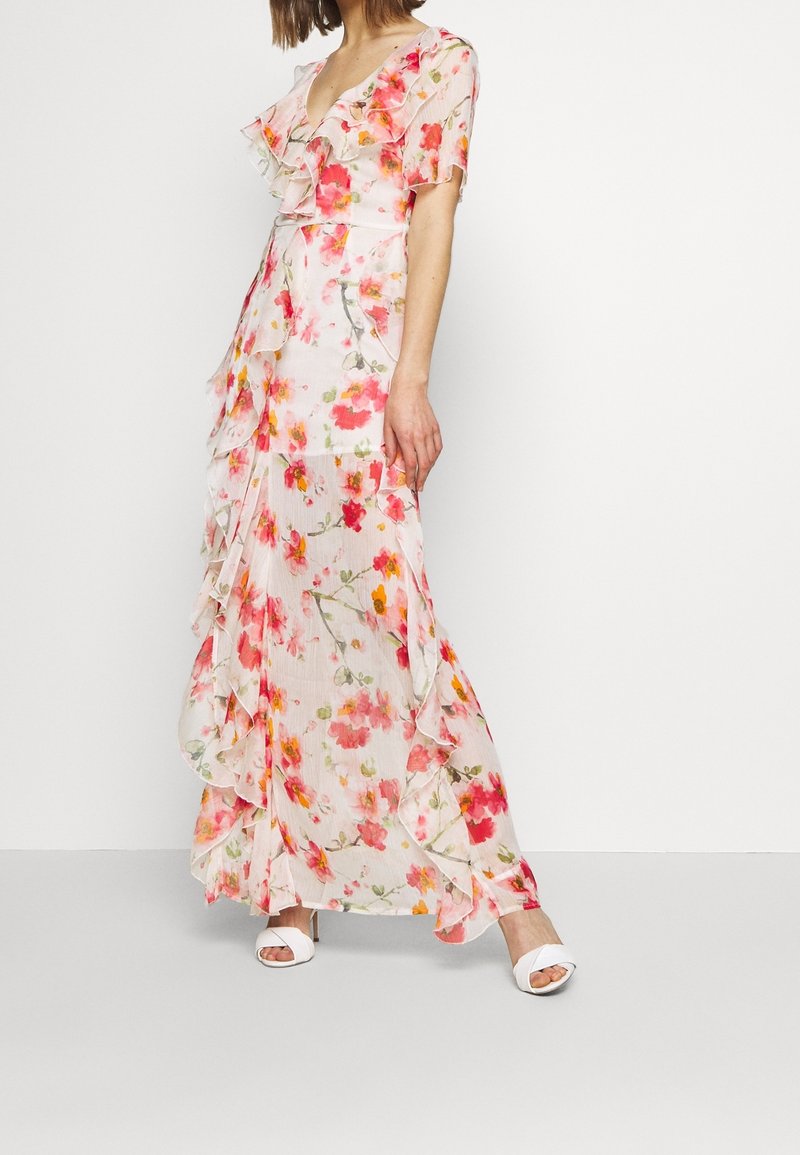 Bloemen maxi-jurk van doorschijnende, lichte stof met roze, oranje en groene bloemen, ruches langs de halslijn en zoom, en een vloeiende silhouet.