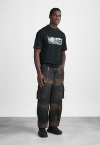 Musta t-paita graafisella printillä, yhdistettynä väljiin, cargo-tyylisiin housuihin, joissa on ruskea sitruunaruokokuvio. Päällä mustat lenkkarit.