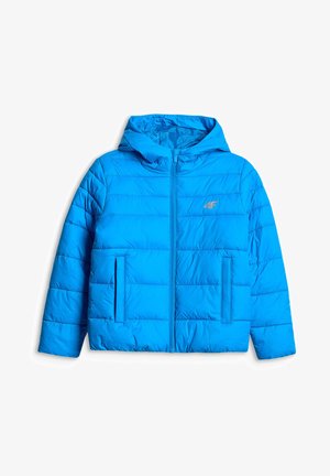 Garçon debout les mains dans les poches portant une doudoune bleue à capuche, une chemise blanche, un pantalon cargo gris et des baskets blanches sur un fond gris uni.