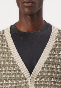 Textureret grøn og beige cardigan med V-hals, ribbede kanter og en enkelt knaplukning over en sort T-shirt med rund hals.