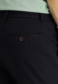 Zwarte broek van gestructureerde stof, met een achterzak met een bruine knop en een gestikte accent boven de zak.