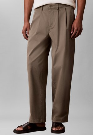 Mężczyzna noszący luźne, jasnobrązowe spodnie z plisami, połączone z ciemnobrązowymi sandałami z odkrytymi palcami, stojący na szarym tle.