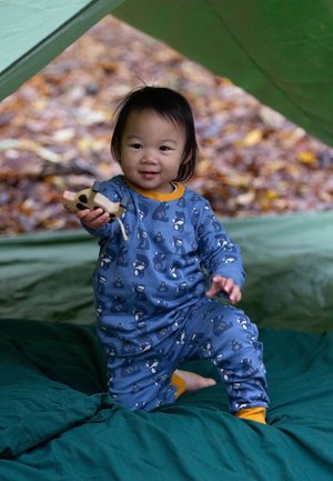 Blaue Schlafanzugset mit Tiermustern, gelben Akzenten, aus weichem Stoff; Kind hält ein Holzspielzeug, sitzt auf grünem Bettzeug in einem Zelt.