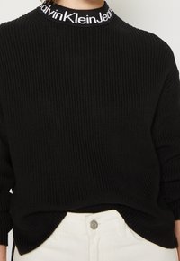 Pull en maille noire avec le texte blanc "Calvin Klein Jeans" sur le col, porté rentré dans un pantalon blanc taille haute avec un bouton.