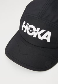 Svart caps med hvit "HOKA"-logo, laget av teksturert stoff, med en strukturert brem og sydd detaljer på toppen.
