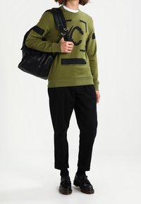 Olivově zelená mikina s černými akcenty a logem, doplněná černými zúženými kalhotami a černými koženými botami, nesoucí černou tašku.