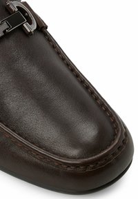 Brun læder loafers med en glat overflade, detaljeret syning og en metallisk detalje på remmen. Rund tå og slip-on design.
