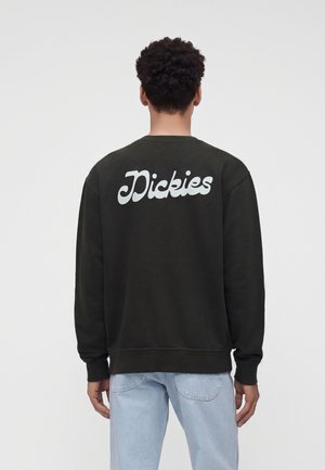 Person med kort krøllet hår iført en sort sweatshirt med hvid "Dickies" tekst på ryggen og lyseblå jeans, stående med ryggen til.