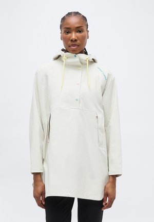 Γυναίκα με πλεξουδάκια στα μαλλιά, φορώντας ανοιχτό μπεζ αδιάβροχο με κουκούλα, παστέλ κουμπιά και κίτρινα κορδόνια, στέκεται μπροστά σε λευκό φόντο.