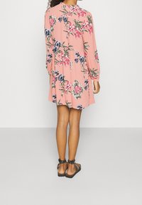 Vaaleanpunainen kukkamekko pitkillä hihoilla, jossa on rento malli ja pyöreä pääntie. Nasta-sandaalit täydentävät asua.