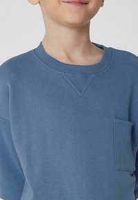 Modrá bavlněná mikina s kulatým výstřihem, krátkým rukávem a kapsou na prsou. Obsahuje žebrovaný límec a detaily na ramenních švech.