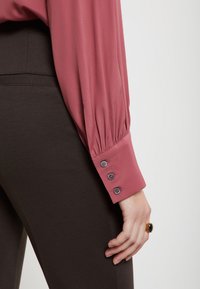 Ružová blúzka s plisovanými dlhými rukávmi a detailom so štyrmi gombíkmi na manžetách, skombinovaná s tmavohnedými priliehavými nohavicami. Celkový hladký vzhľad.