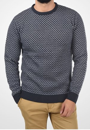 Homme portant un pull bleu marine et blanc à motifs avec des poignets et un ourlet côtelés, associé à un pantalon marron clair, les mains dans les poches.