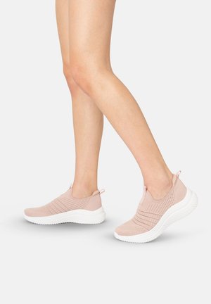 Benen met lichtroze gebreide slip-on sneakers met witte zolen tegen een effen lichte achtergrond.
