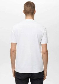 Un homme vu de dos portant un t-shirt blanc à manches courtes et un pantalon noir sur fond blanc.