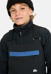 Teenage dreng med bøjler iført en sort hue, sort zip-up jakke og sorte overalls med blå lomme og Quiksilver-logo.