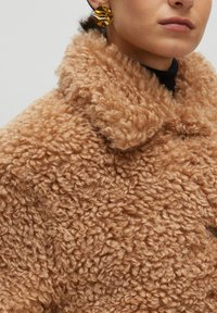 Beige, strukturierter Mantel aus flauschigem Material mit einem hohen Kragen. Er weist ein weiches, lockiges Erscheinungsbild und sichtbare Nähdetails auf. Goldene Ohrringe runden den Look ab.