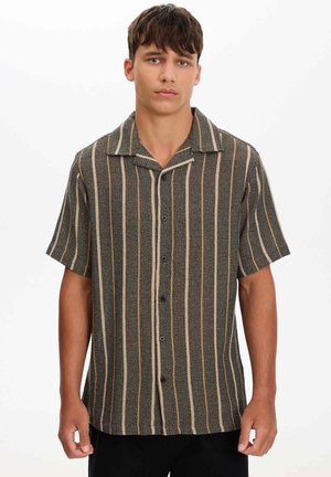 Joven con camisa de manga corta abotonada con rayas verticales beige y naranja sobre fondo oscuro, de pie frente a un fondo blanco.