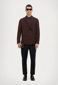 Rjav dvojno zapet blazer s črnimi gumbi, kombiniran s temno srajco in črnimi hlačami. Dodatki vključujejo sončna očala in majhno črno denarnico.