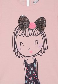 Lichtroze shirt met een cartoonmeisje met een zwart en roze pailletten strik, florale details op een donkere jurk en omrande gelaatstrekken.