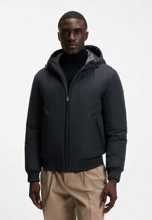Mand iført en sort hættetrøje dunjakke over en sort sweater og beige bukser, stående mod en ensfarvet hvid baggrund.