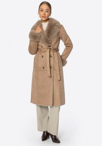 Abrigo de ante sintético beige con un gran cuello de piel, diseño de doble botonadura, cinturón en la cintura y bolsillos laterales, llevado sobre pantalones de color claro.