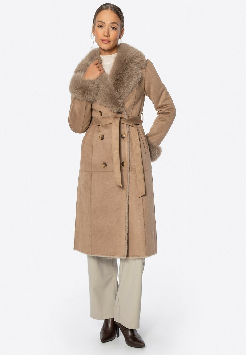 Abrigo de ante sintético beige con un gran cuello de piel, diseño de doble botonadura, cinturón en la cintura y bolsillos laterales, llevado sobre pantalones de color claro.