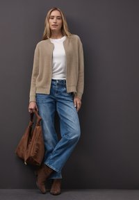 Cardigan beige duveteux porté sur une chemise blanche, associé à un jean large bleu clair. Porte des bottines en daim marron et tient un sac marron.
