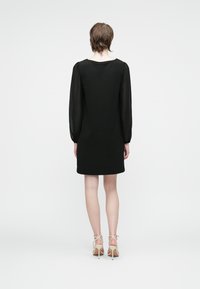 Woman facing away wearing a black long-sleeve dress with sheer sleeves and beige high-heeled sandals on a white background.