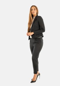 Veste noire sur mesure en tissu texturé, assortie à un pantalon slim-fit. Portée avec un haut blanc et des talons hauts noirs.