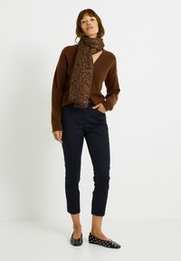 Suéter marrón tejido con cuello en V, combinado con pantalones negros recortados y zapatos planos negros adornados con lunares blancos. Bufanda marrón con estampado envuelta alrededor del cuello.