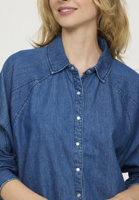Femme souriante portant une chemise en denim bleue à boutons, avec des manches retroussées et des cheveux blonds lâches.
