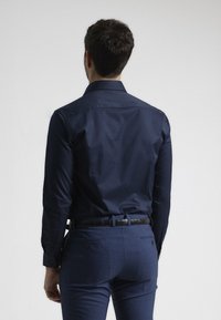 Camisa de manga larga azul marino con cuello estándar. Textura suave, diseño ajustado, mostrada por la parte de atrás. Lleva pantalones oscuros y un cinturón.