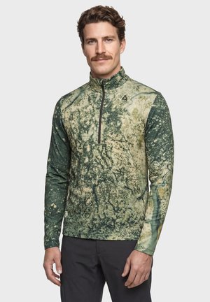 Mann mit braunen Haaren und Schnurrbart, der einen grün-beige gemusterten Langarm-Pullover mit Frontreißverschluss trägt, steht vor einem schlichten Hintergrund.