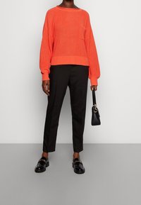 Jasně oranžový pletený svetr s dlouhými rukávy a kapsou, doplněný černými šitými kalhotami a černými mokasínami, držící malou černou kabelku.