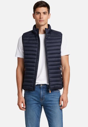 Giovane con gilet imbottito blu navy sopra una t-shirt bianca e jeans blu, in piedi davanti a uno sfondo chiaro e semplice.