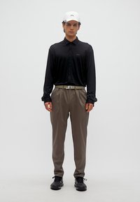 Άνδρας που φοράει μαύρο πουκάμισο με μακριά μανίκια, μπεζ πιετέ παντελόνια, λευκό καπέλο, μαύρα παπούτσια και μπεζ ζώνη, στέκεται μπροστά σε λευκό απλό φόντο.