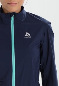 Veste légère bleu marine avec un col montant, une fermeture éclair frontale turquoise et un logo argenté sur la poitrine. Texture lisse, design moderne.