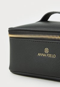 Neceser de maquillaje negro con textura, con cremallera dorada y logotipo. Forma rectangular compacta con un asa superior para un fácil transporte.