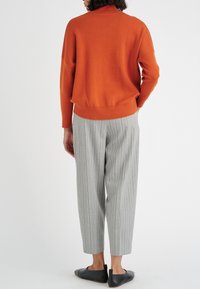 Person som står med ryggen vänd, iklädd en orange tröja, grå randiga byxor och svarta slipskor mot en vit bakgrund.