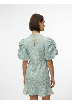 Femme portant une robe fleurie vert menthe avec des manches bouffantes et une petite ouverture en forme de trou à l'arrière, debout de dos sur un fond uni.