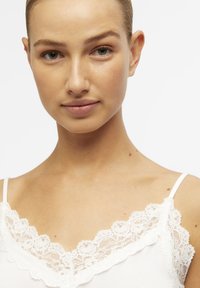 Hvid top med blonderkant ved halsudskæringen, med tynde stropper og en glat overflade, der fremhæver et delikat blomstermønster i blonderne.