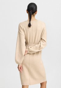 Beige Strickkleid mit langen Ärmeln, lässiger Passform und einem gebundenen Bund. Das Material wirkt weich, mit gerippten Bündchen und einem nahtlosen Rückendesign.
