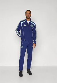 Tumesinine dressipluus helesiniste detailidega, millel on lukuga jakk ja kitsenevad püksid. Mõlemal tükil on Adidase logo ja embleem.