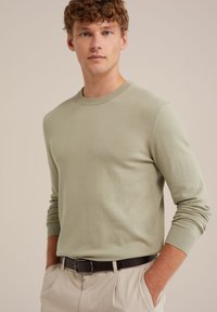 Suéter de punto verde claro con cuello redondo, mangas largas y puños acanalados, combinado con pantalones beige y un cinturón marrón.