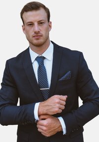 Costume bleu marine avec une chemise blanche et une cravate à pois. Comprend un mouchoir de poche, des boutons de manchette et une pince à cravate en argent. Coupe ajustée.