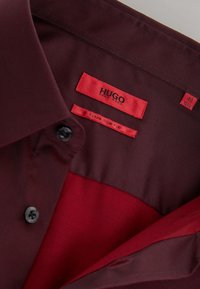 Camisa de vestir burdeos con una textura suave, con un botón negro, etiquetas rojas que dicen "HUGO" y "Extra Slim Fit", y etiqueta de tamaño 40.
