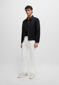 Uomo in piedi che indossa una giacca a zip nera, una maglietta nera, pantaloni bianchi e sneakers bianche su uno sfondo grigio chiaro uniforme.